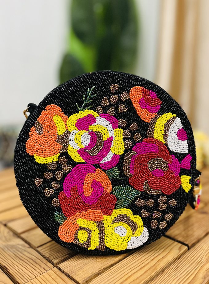 A black circular hand-beaded shoulder bag with intricate floral patterns and a chain strap, resting on a sleek, glossy